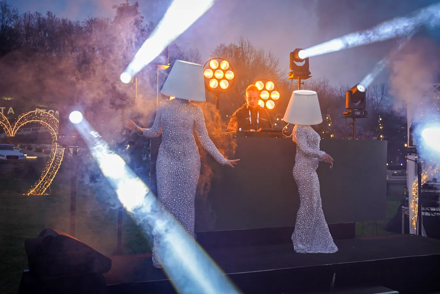 DJ in consolle e due performer con abiti di paillettes, tra fasci di luce e fumo